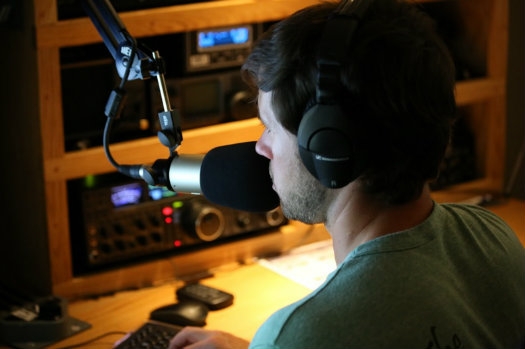 A man speaking into a microphone next to a radio station set up.